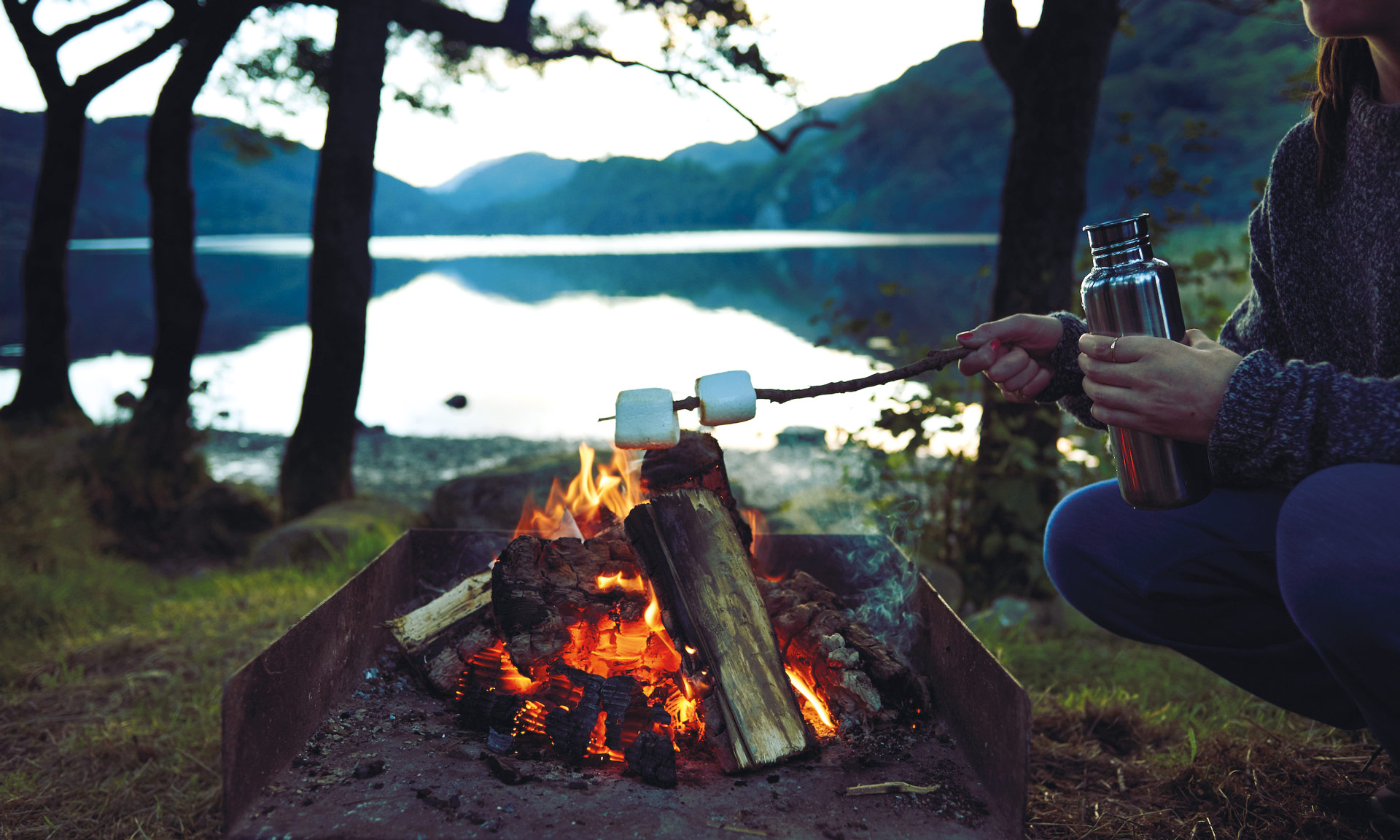 A camper tasting marshmallows above an open fire holding a flask with a lake in the background