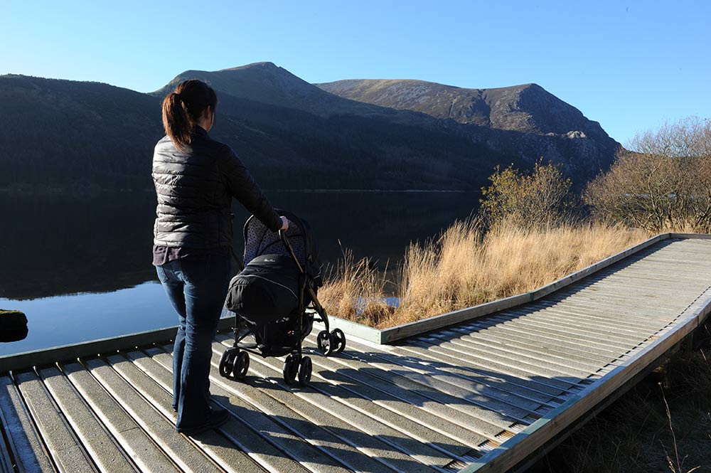 A person standing with a pram on the Llyn Cwellyn Boardwalk, enjoying a sunny day.