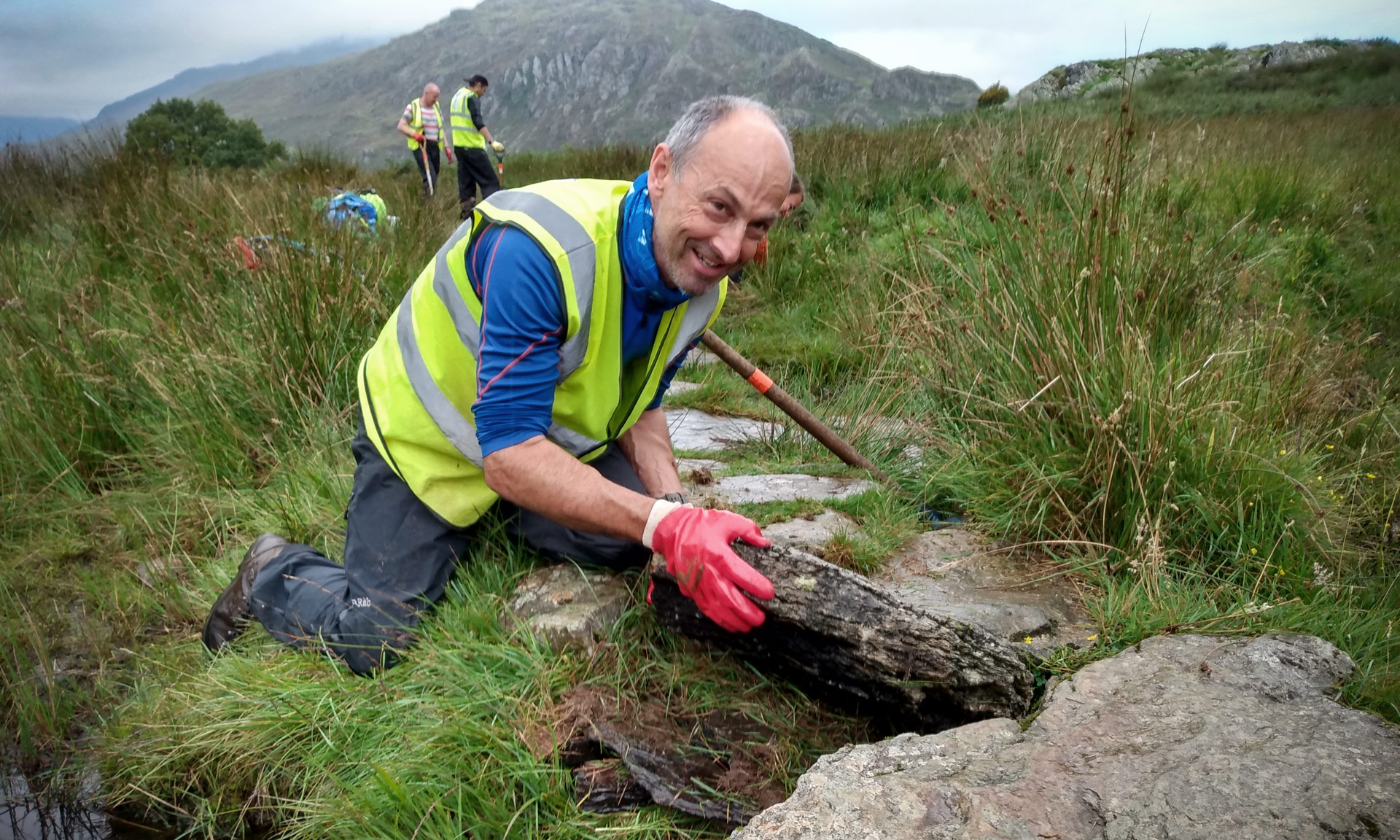 A volunteer works on a stone path, smiling! Wearing red gloves and a high vis jackets