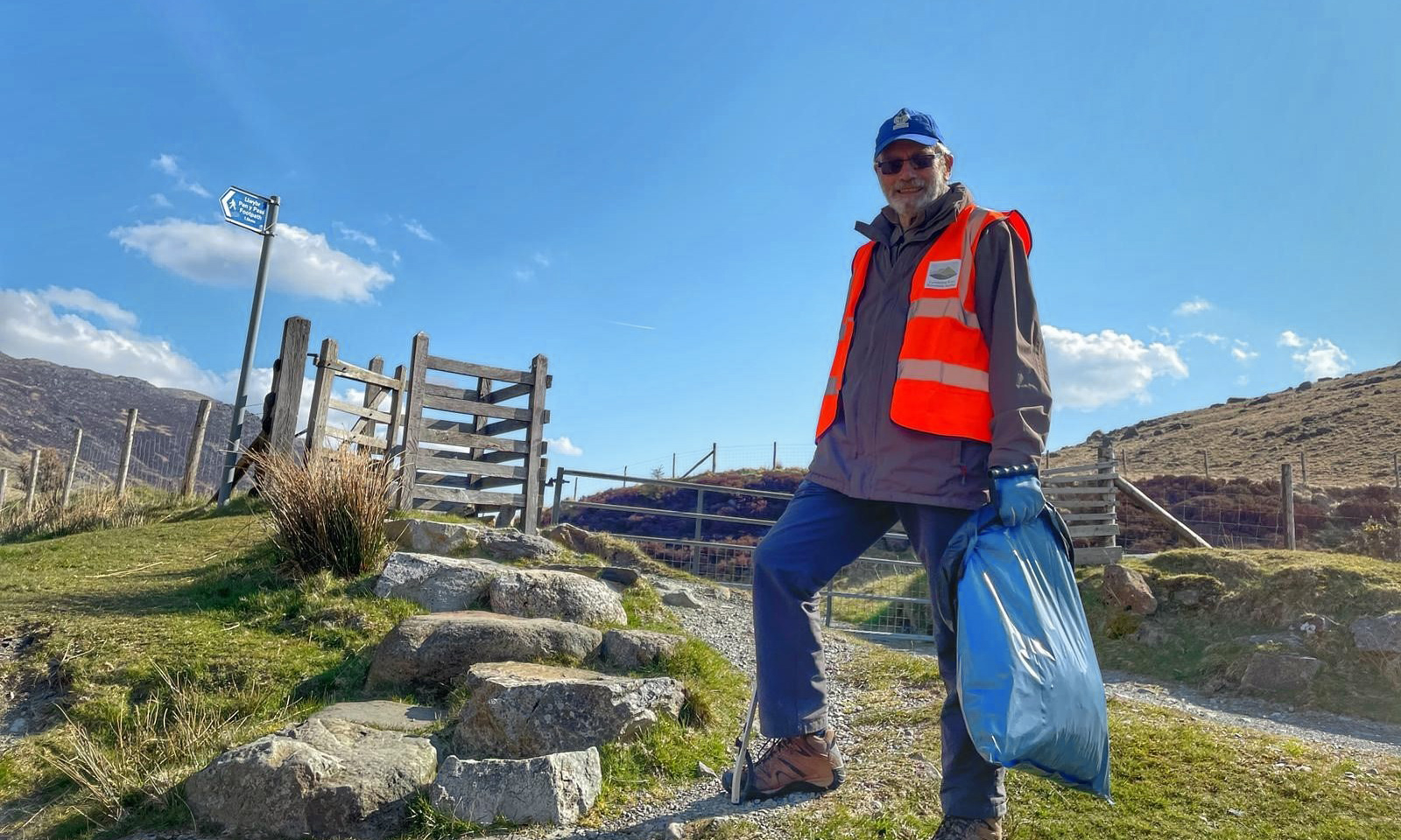 A volunteer wearing a Snowdonia Society high-visibility jacket stands holding a litter picker and a bin bag, smiling happily against a scenic backdrop.