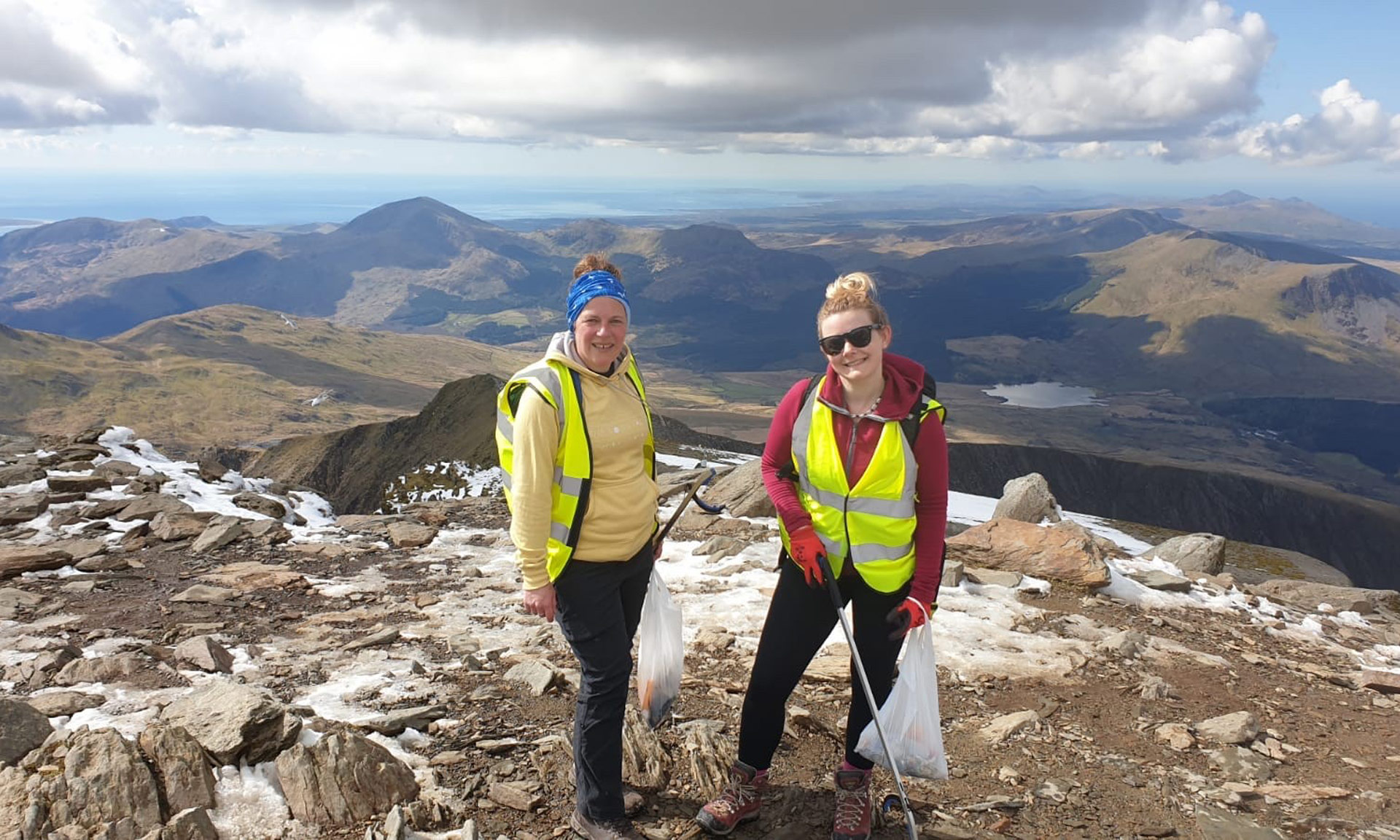 Two of our Volunteer Wardens smiling with the mountains in the background, wearing high vis jackets. A bit of snow is visible on the floor.