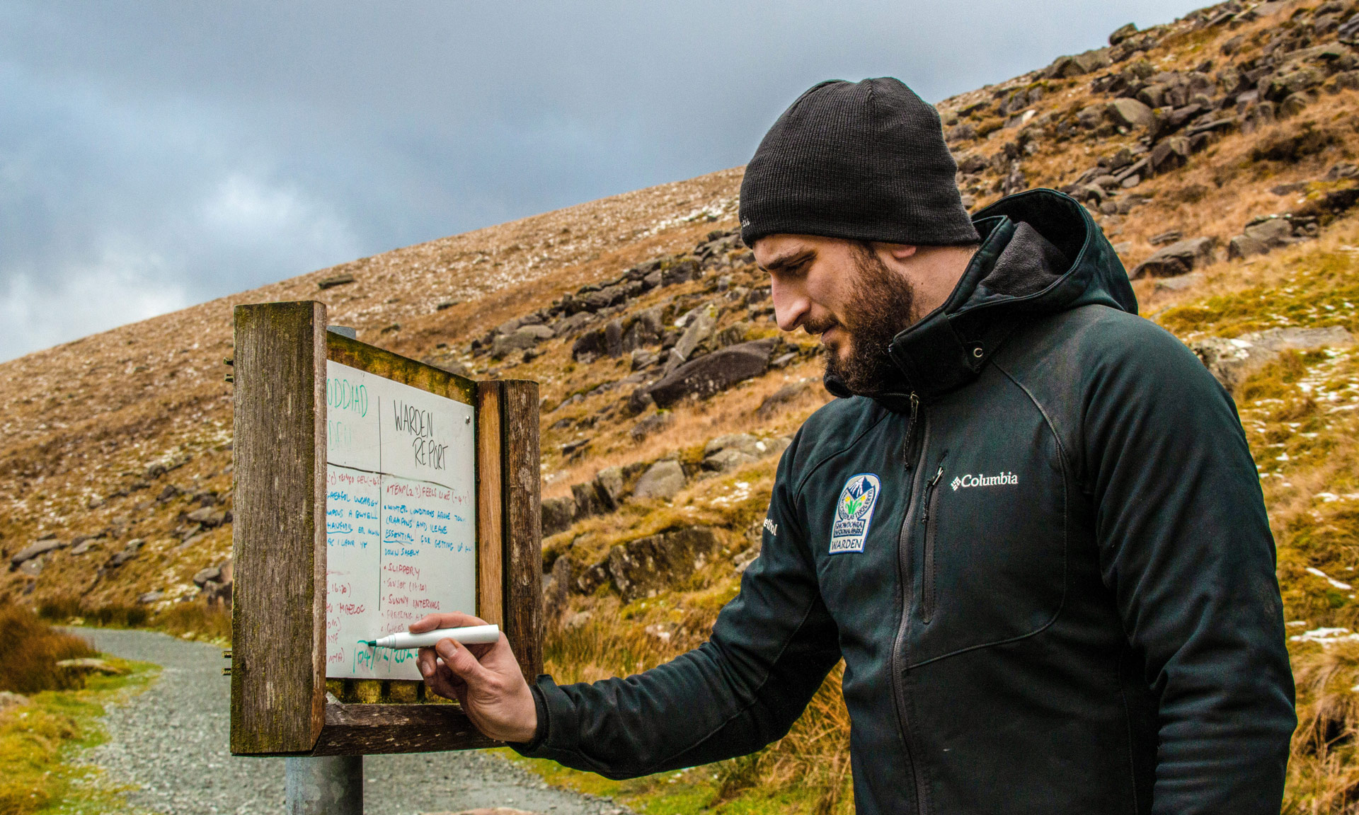 A Warden in Eryri National Park clothing writes the weather forecast on a whiteboard using a green marker.