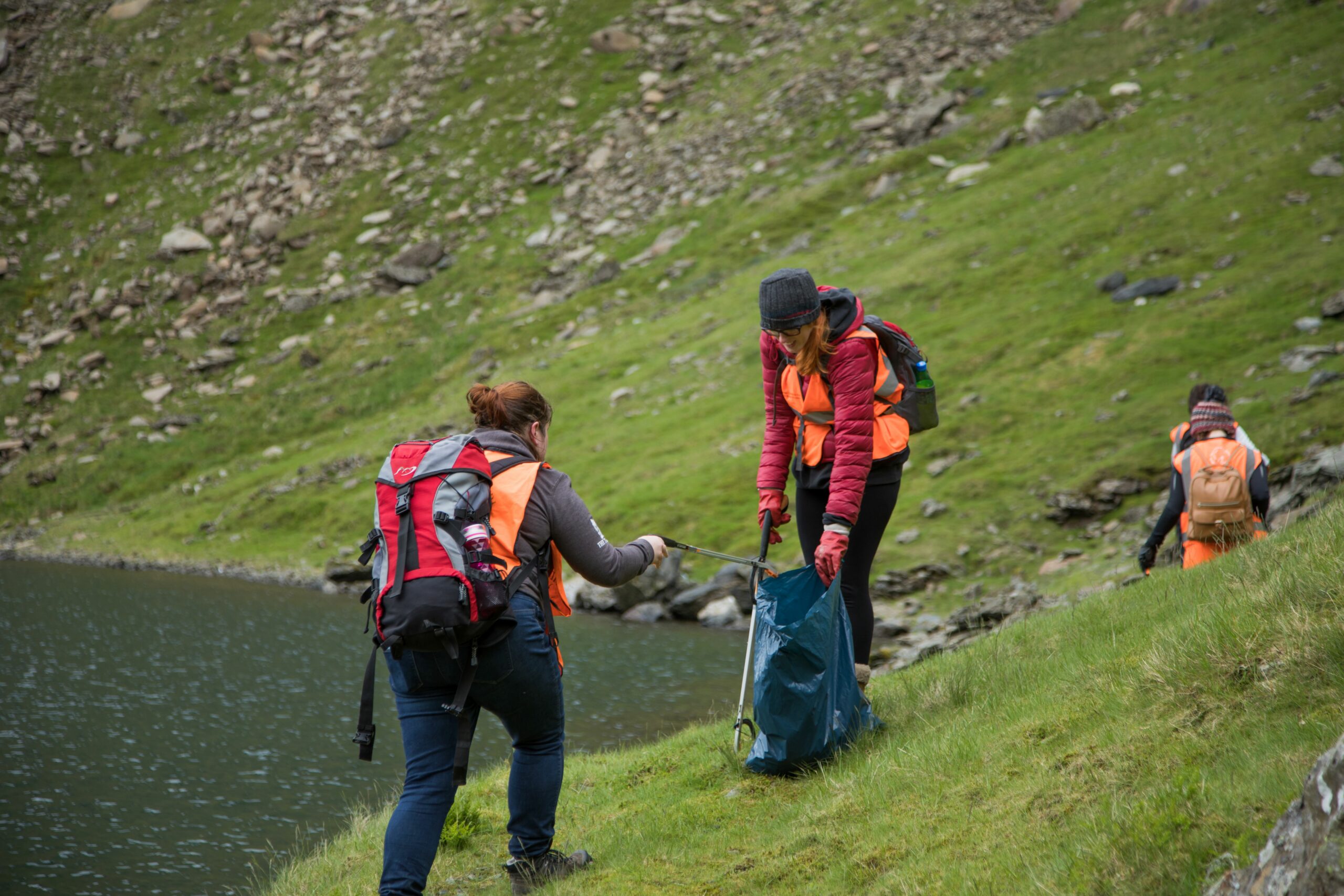 Two individuals from Caru Eryri are engaged in litter picking on Yr Wyddfa. They are equipped with litter bags and grabbers, surrounded by a scenic mountainous landscape, symbolising their commitment to maintaining the natural beauty of the area.